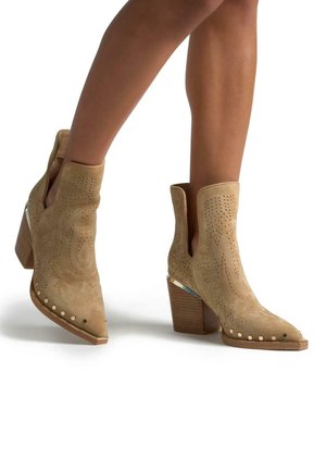 Tan-farbene Wildleder-Ankle-Boots mit spitzer Zehenform, perforiertem Design, dekorativen Nietenakzenten und einem gestapelten Holzabsatz.