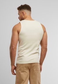 Beige ribbet tanktop med en tætsiddende design. Personen har kort krøllet hår og har tanbukser på. Synlige rygmuskler og stoftekstur.