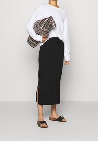 Jupe maxi noire avec des fentes sur les côtés, associée à un haut blanc à manches longues et des sandales noires. Porte un sac à main en toile à carreaux marron.