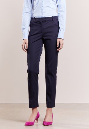 Femme portant un pantalon bleu marine sur mesure, une chemise boutonnée bleu clair et des chaussures à talons hauts rose vif, debout sur un sol neutre.