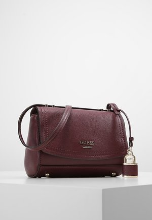 Donker bordeauxrode leren Guess-handtas met schouderriem, zilveren logo en decoratieve hangslotbedel op een witte ondergrond.