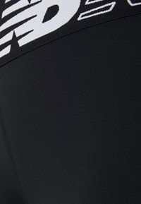 Černé legíny s vysokým pasem a texturovaným pasem, které mají bílé branding. Materiál je hladký s matným povrchem.