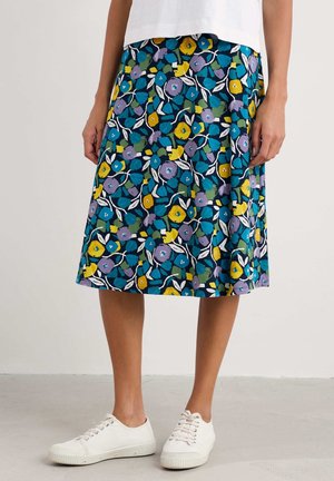 Femme portant une jupe longueur genou avec des motifs floraux bleus, jaunes et violets, un haut blanc et des baskets blanches, debout sur un sol uni.