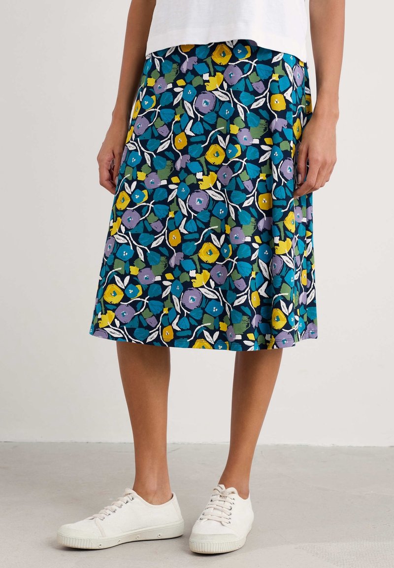 Femme portant une jupe longueur genou avec des motifs floraux bleus, jaunes et violets, un haut blanc et des baskets blanches, debout sur un sol uni.