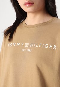 Braunes Baumwoll-T-Shirt mit Rundhalsausschnitt, auf der Vorderseite mit dem weißen Text "TOMMY HILFIGER EST. 1985" bedruckt. Glatte Textur.