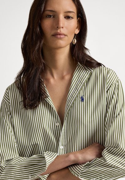 Γυναίκα με μακριά σκούρα μαλλιά που φοράει ριγέ πουκάμισο με κουμπιά σε πράσινο και λευκό, με μπλε λογότυπο, σκουλαρίκι κοχύλι, σταυρωμένα χέρια, ουδέτερο φόντο.