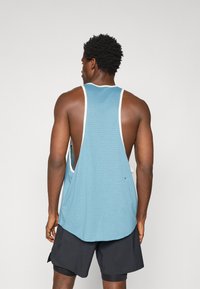 Top desportivo azul em malha, com detalhes brancos e aberturas profundas nas axilas. Combinado com calções pretos. Vista traseira mostrando todos os detalhes do design.