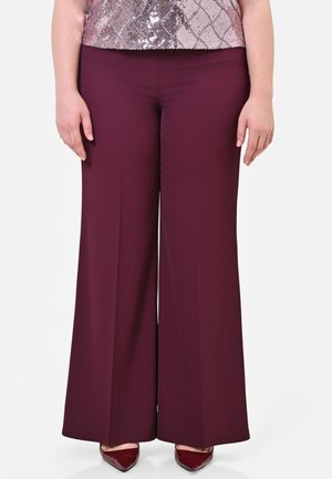 Persona che indossa pantaloni larghi color amaranto, top rosa con paillettes e tacchi color amaranto dalla punta appuntita, in piedi con le mani rilassate ai lati.