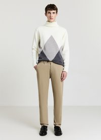 Strick-Turtleneck-Pullover in Creme mit grauen Rautenmuster, kombiniert mit beigen Hosen und schwarzen Schuhen, in einer lässigen Passform.