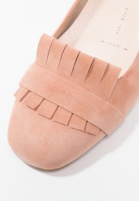 Gros plan d’une chaussure plate en daim rose avec une frange décorative et une sangle horizontale au niveau des orteils.
