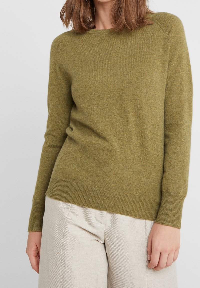 Femme portant un pull à manches longues vert olive et un pantalon beige clair, debout avec une main le long du corps sur un fond uni.