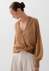 Suéter marrón de punto texturizado con escote en V profundo y puños acanalados, combinado con pantalones de talle alto en beige con pliegues y detalle de botón.