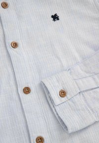 Jasnoniebieska, fakturowana koszula z zapinaniem na guziki z przodu, drewnianymi guzikami i małym ciemnym logo na piersi. Posiada detal w postaci złożonego mankietu.