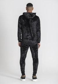 Svart sammetsträningsdräkt med huva, med mjuk textur och slim fit. Bärs med mönstrade sneakers, vilket visar en elegant bakvy.