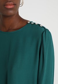 Detailní záběr na rameno tmavé pleti ženy, která má na sobě azurovou blůzu s dekorativními knoflíky a nařasenými rukávy.