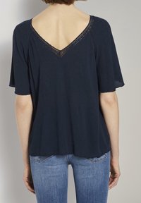 Donna che indossa un top blu navy a maniche corte con scollatura a V bordata di pizzo, abbinato a jeans di denim blu chiaro, in piedi contro un muro semplice.
