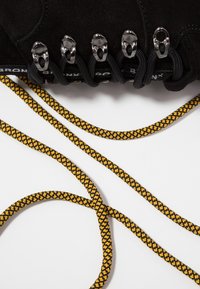 Œillets de bottes en suède noir avec des lacets noirs partiellement noués, et une corde d'escalade jaune et noire posée sur une surface blanche.