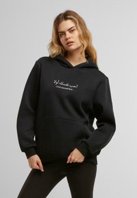 Felpa nera con cappuccio realizzata in tessuto morbido, dotata di tasca frontale e coulisse, con testo bianco in arabo e inglese.