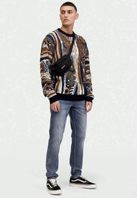 Maglione colorato a motivo nelle tonalità di marrone, blu e bianco, abbinato a jeans azzurri chiari e sneakers nere. Inclusa borsa a tracolla nera.