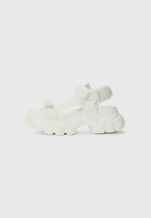 Sandalia blanca con parte superior de malla, suela gruesa con diseño ondulado, correas ajustables y logo en la correa.