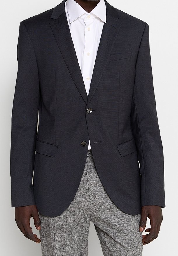 Homme portant un blazer noir sur une chemise blanche et un pantalon gris à motifs, debout devant un fond uni.