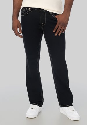 Uomo che indossa jeans neri con cuciture bianche e sneakers bianche, in piedi con una mano in tasca davanti a uno sfondo semplice.