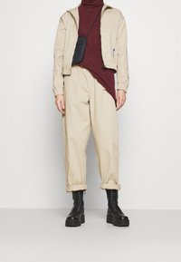 Beige lightweight jacket over a burgundy turtleneck, paired with beige rolled-up trousers and black lace-up boots. Small black crossbody bag.