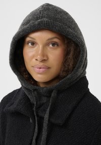 Pardoseală texturată de culoare neagră cu o glugă tricotată de culoare gri. Gluga dispune de șnururi; materialul pare moale și confortabil, cu o închidere cu nasture la guler.