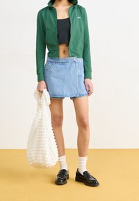 Grön zip-up hoodie över en svart cropped topp, jeanskjol med omlott, svarta loafers, vit stickad väska och vita ankelsockor på ett gult golv.