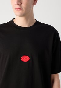 Black cotton t-shirt featuring a raised red rubber logo in an oval shape at the center, with a classic round neckline and short sleeves.