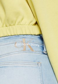 Sudadera amarilla clara con dobladillo elástico, combinada con jeans de mezclilla azul claro. Los jeans presentan la marca "Calvin Klein Jeans" en hilo beige.