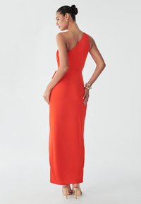 Model in een levendig rode, een-schouder jurk, met de rug naar de camera, die een slanke silhouette en beige hoge hakken toont, tegen een neutrale achtergrond.