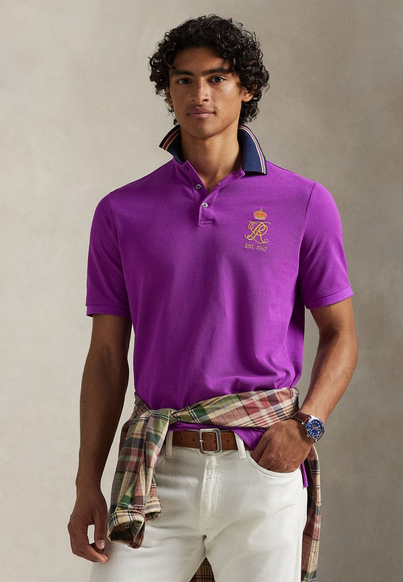 Μωβ πόλο μπλούζα με σκούρο μπλε γιακά που έχει πολύχρωμες ρίγες. Χρυσαφί κεντημένο λογότυπο στο αριστερό στήθος. Συνδυάζεται με πουκάμισο καρό δεμένο στη μέση.