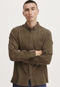 Olivově zelená košile na knoflíky s kapsou na prsou, dlouhými rukávy a volným střihem. Tkanina vypadá měkce s mírným leskem.