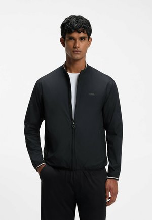 Junger Mann mit lockigem schwarzen Haar, der eine schwarze Jacke mit Reißverschluss über einem weißen Hemd trägt, steht vor einem schlichten hellen Hintergrund.