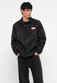 Μαύρο μπουφάν από λείο ύφασμα με κολάρο, κουμπιά με κουμπωτή σύνδεση, ελαστικούς καρπούς και κόκκινο λογότυπο στο στήθος.