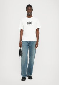 Hvit t-skjorte med svart "MK" logo, kombinert med lyseblå jeans. Modellen holder en svart veske. Enkelt design, kortermet og avslappet passform.