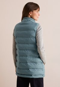 Gedämpfte Türkis-Weste mit hohem Kragen, horizontaler Steppung und seitlichen Taschen, getragen über einem hellgrauen Pullover.