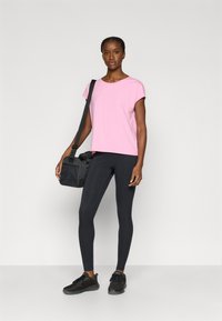 Ροζ πουκάμισο με κοντά μανίκια, μαύρο κολάν και μαύρα αθλητικά παπούτσια. Το μοντέλο κρατά μια μαύρη τσάντα ώμου, στέκεται μπροστά σε ουδέτερο φόντο.