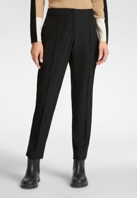 Schwarze maßgeschnittene Hosen mit vorderen Falten und einem Knopfverschluss. Glattes Material, gerade Passform, schmal zulaufende Beine, kombiniert mit schwarzen Knöchelstiefeln.