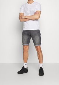 Camiseta blanca con diseño gráfico, pantalones cortos de mezclilla gris con dobladillos enrollados y zapatillas negras con calcetines blancos. El modelo está de pie con los brazos cruzados.