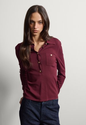 Mujer con cabello largo y oscuro que lleva una camisa burdeos de manga larga abotonada y pantalones azul oscuro sobre un fondo claro y liso.