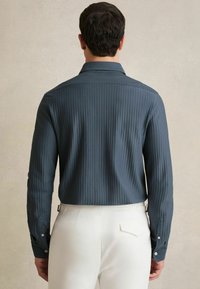 Camisa de manga larga en tejido azul oscuro texturizado, con rayas verticales, un cuello clásico y puños abotonados, combinada con pantalones de color blanco roto.