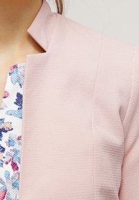 Lichtroze blazer met een getextureerde stof; beschikt over een hoge kraag en een gestructureerd ontwerp. Bloemmotief zichtbaar bij de halslijn.