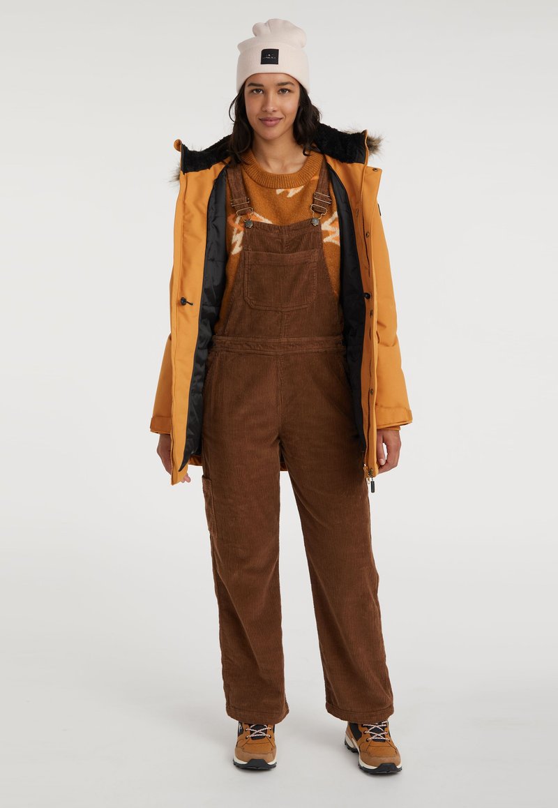 Overoles de pana marrones con tirantes ajustables, un suéter naranja con un patrón y una chaqueta aislante amarilla. Botas de senderismo color beige completan el conjunto.