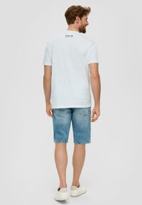 Camiseta de algodón blanca con un logo negro en la parte posterior, combinada con pantalones cortos de mezclilla claros y zapatillas blancas, vista desde la espalda.