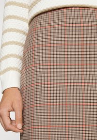 Brun rutig kjol med ett mönster av svarta och röda linjer, tillverkad av texturerad tyg, med en hand som varsamt greppar midjebandet.