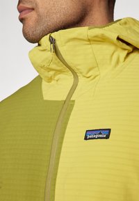 Patagonia jaka zaļā-dzeltenā krāsā ar teksturētu audumu, ar rāvējslēdzēju priekšā un kapuci. Ir logotips kreisajā krūts rajonā.