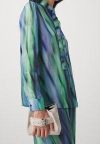 Camicia trasparente con bottoni in gradiente verde e blu, con una consistenza fluida. Abbinata a una borsa leggera e strutturata con dettaglio a fibbia.