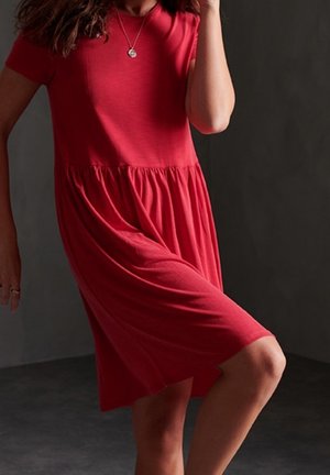 Donna che indossa un abito rosso fluente a maniche corte con vita arricciata, ginocchio sollevato e una collana con ciondolo rotondo su uno sfondo scuro.